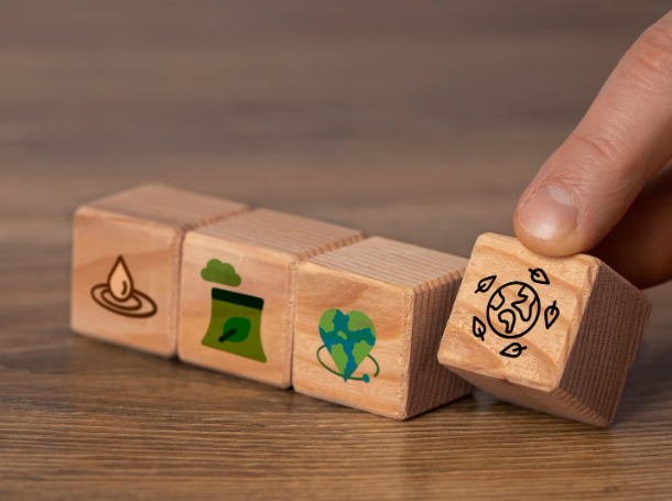 Mano colocando un bloque de madera con un icono del planeta Tierra y hojas verdes junto a otros bloques con símbolos de sostenibilidad, representando acciones para reducir la huella de carbono y proteger el medio ambiente.